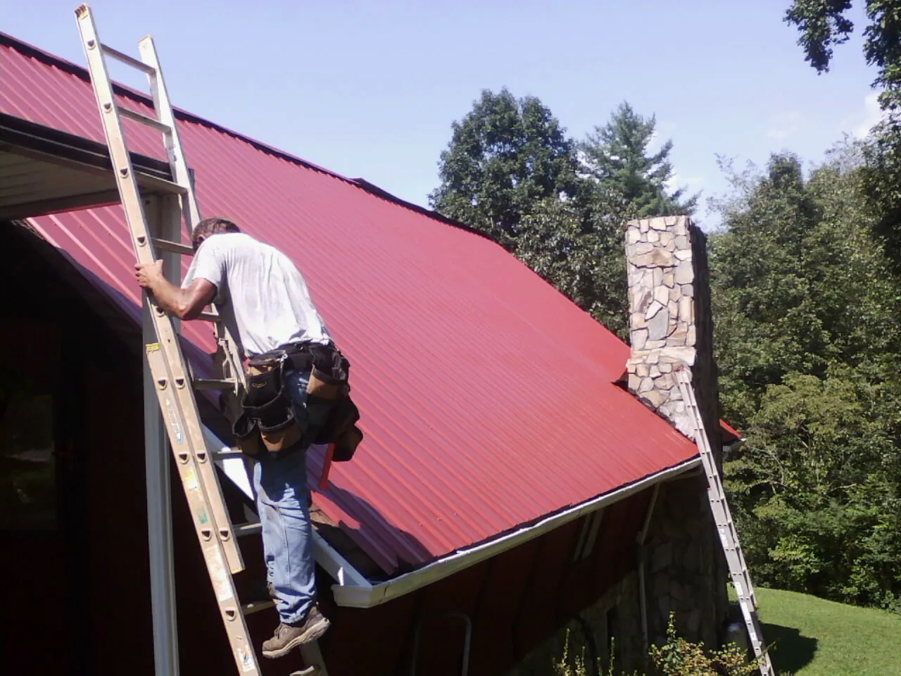 Roofer on a ladder beside a red metal roof and stone chimney — Soffit & Fascia Repair by Roofing Brothers in Southlake